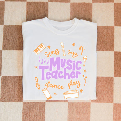 Music Teacher Shirt. A white shirt with the words Music Teacher, Sing, Say, Dance, Play, and drawings of musical instruments.