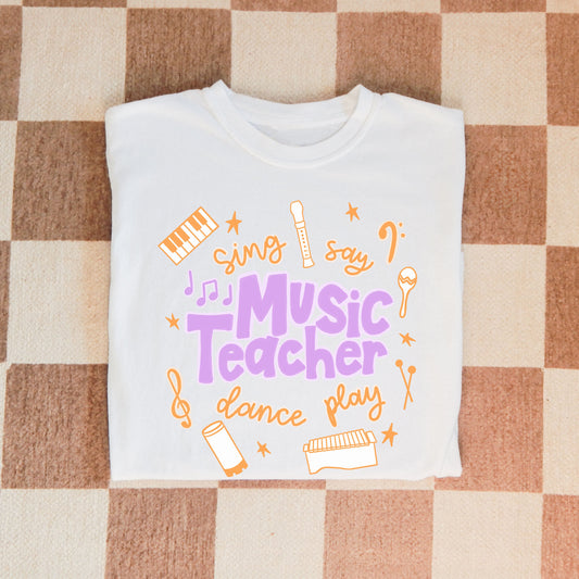 Music Teacher Shirt. A white shirt with the words Music Teacher, Sing, Say, Dance, Play, and drawings of musical instruments.