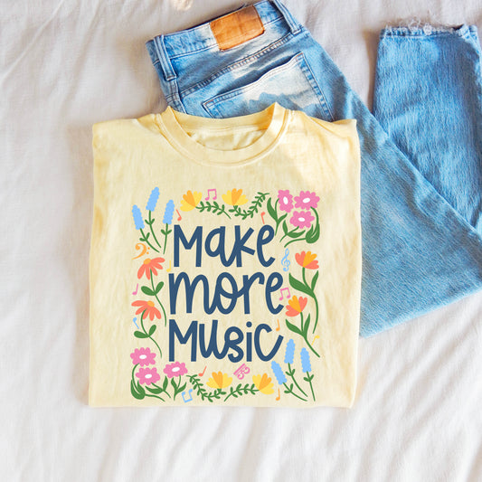 Yellow t-shirt with 'Make more music' text and floral designs on a white surface with blue jeans.